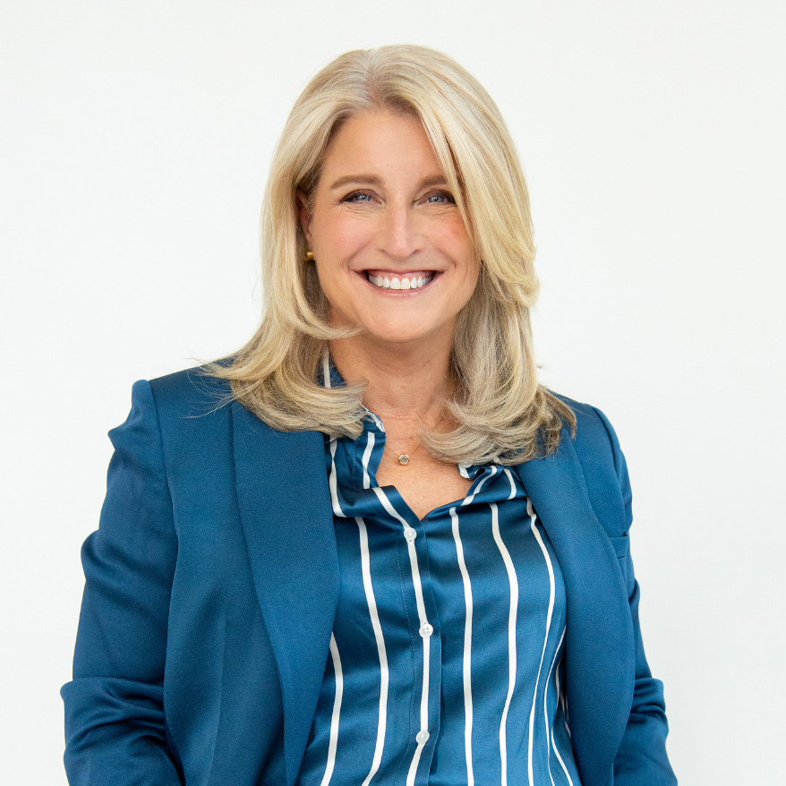 Professional headshot of a smiling blonde woman in a blue blazer and striped shirt against a light background.