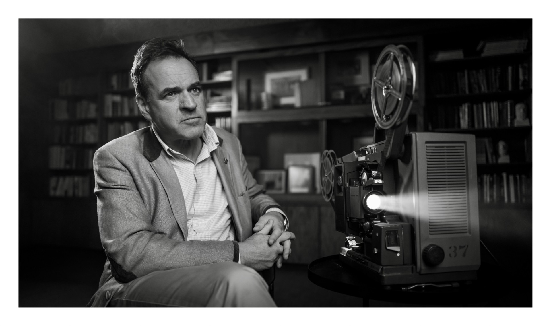 Man in a light blazer sits at a table with a vintage film projector beside him in a study or library setting.