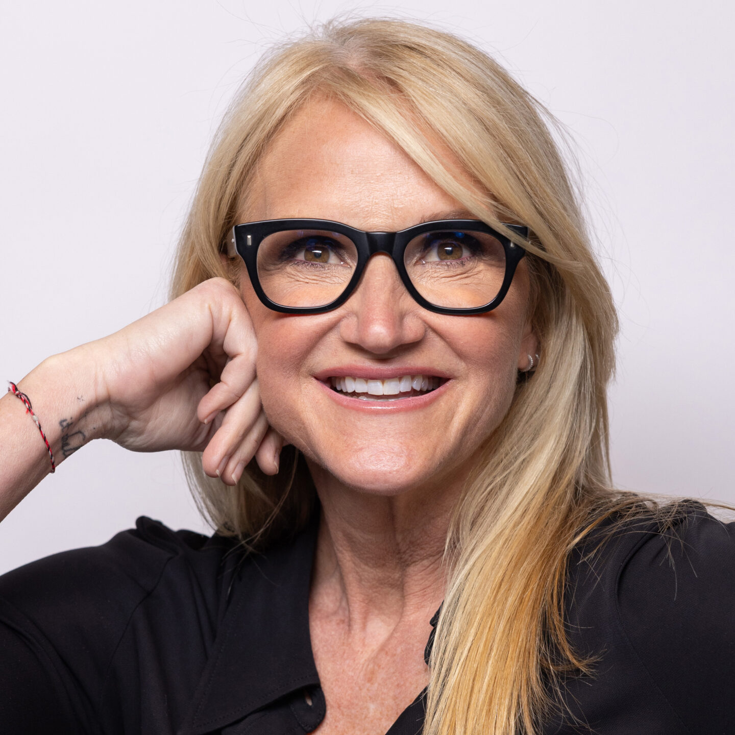 Smiling blonde woman wearing black thick-framed glasses rests her chin on her hand against a light background.