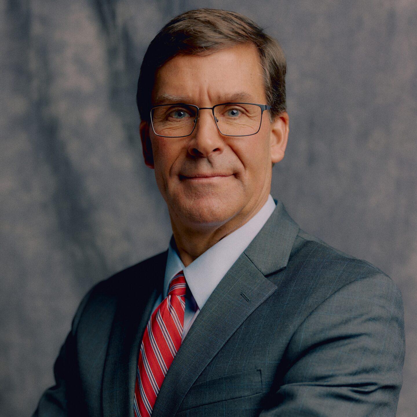 Professional headshot of a middle-aged man in a gray suit and red striped tie, wearing glasses and a slight smile, against a neutral backdrop