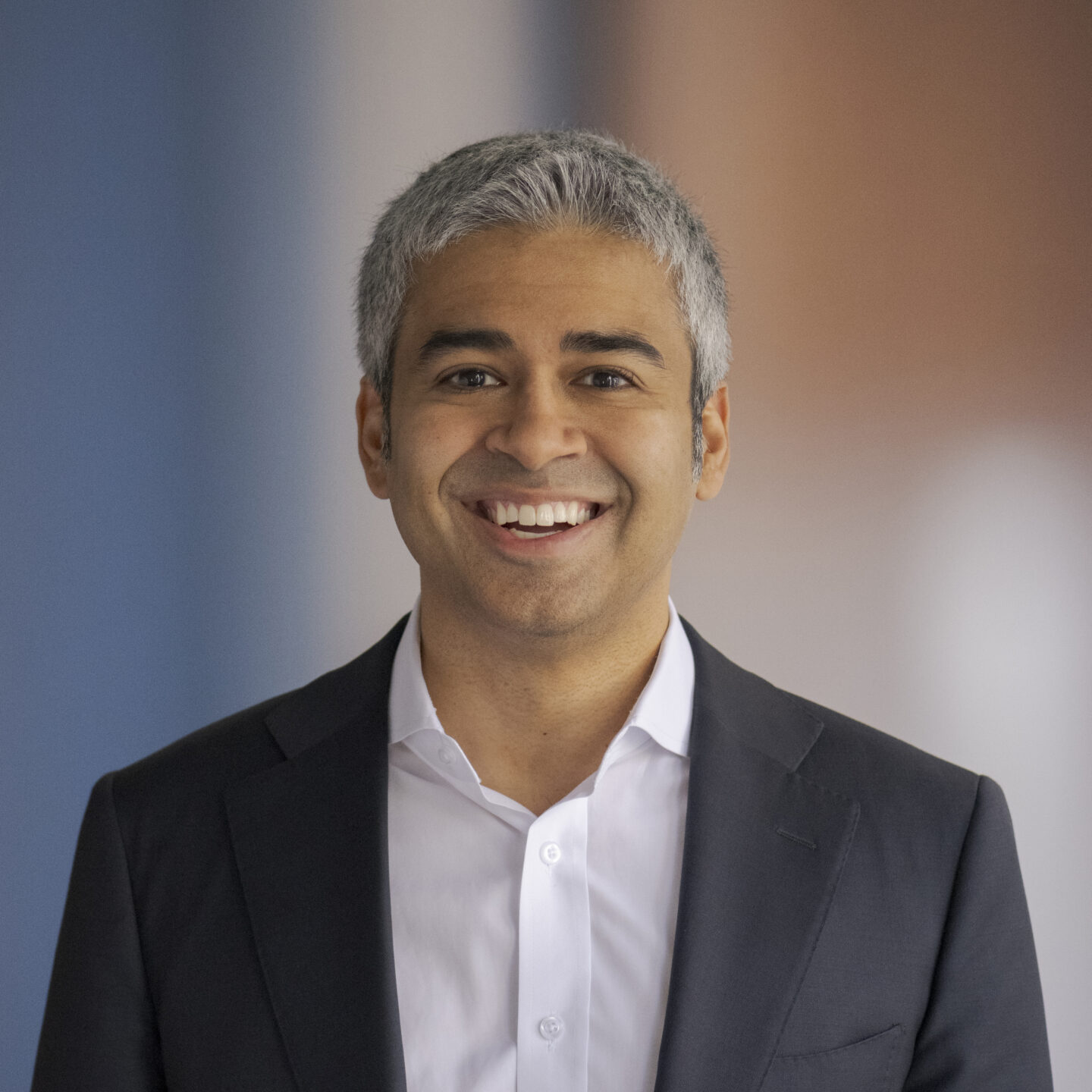 Professional headshot of a man in a dark suit smiling against a blurred blue-tan background.
