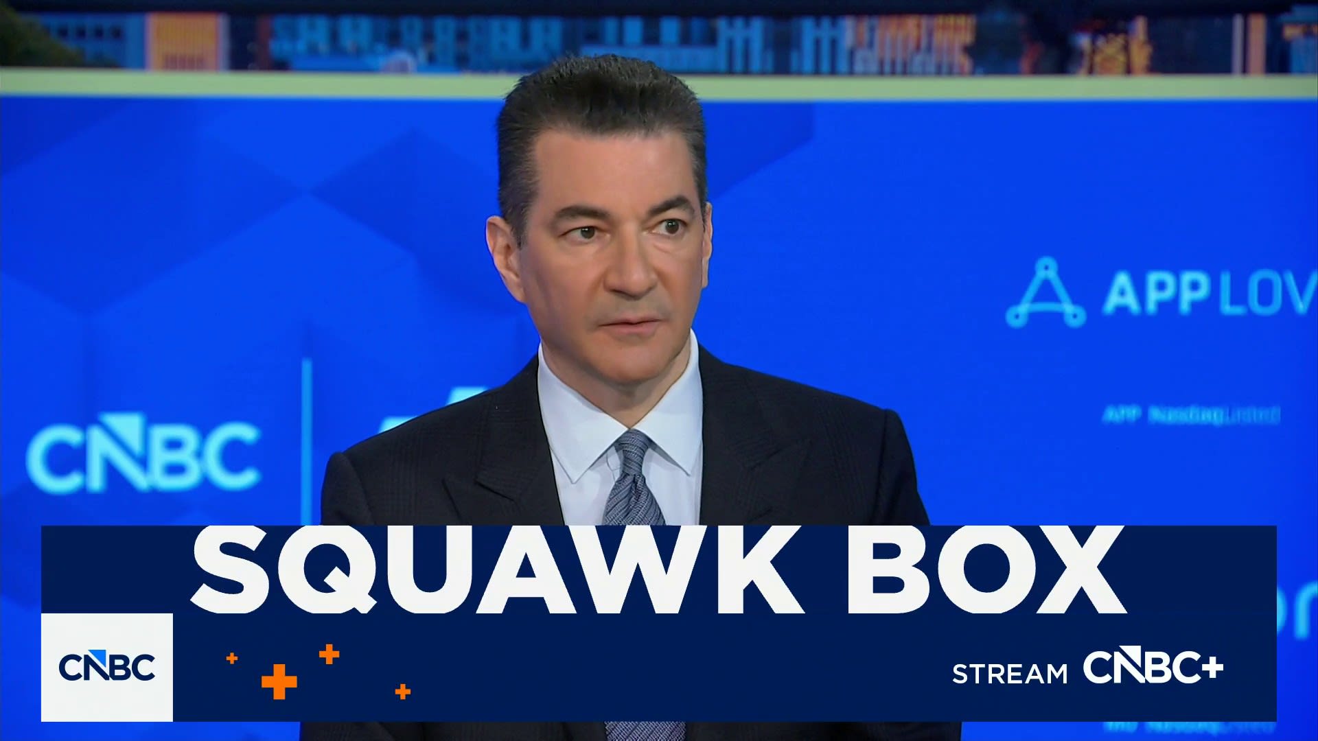 Male news anchor in a dark suit and tie on CNBC's Squawk Box set, with a large 'SQUAWK BOX' banner and CNBC logos.
