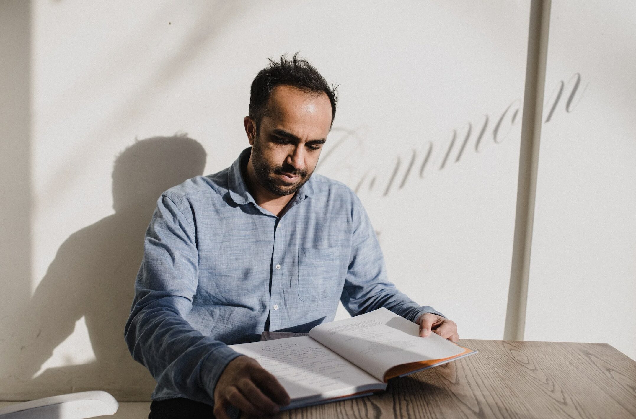 Man in a blue shirt sits at a wooden table, reading an open book with a focused expression as sunlight casts shadows on the wall.