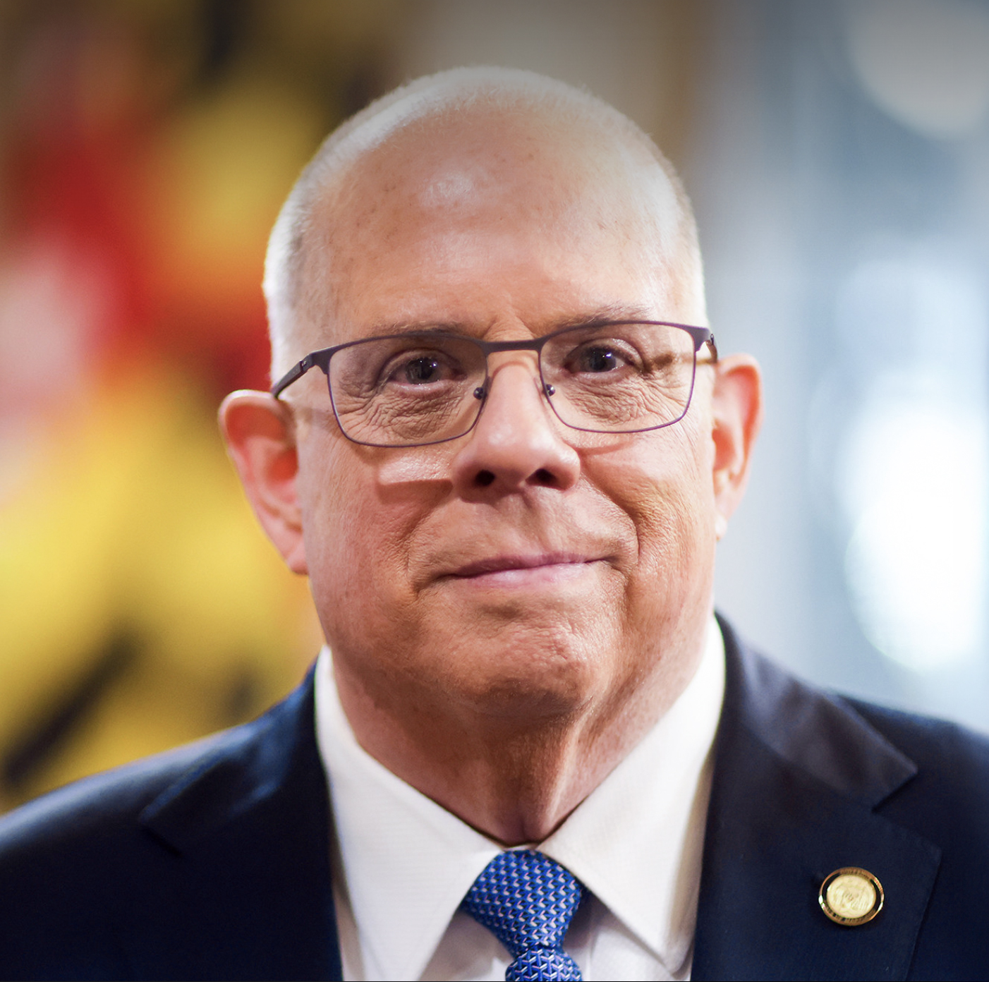 Close-up portrait of a bald man in a dark suit, white shirt, and blue tie wearing glasses.