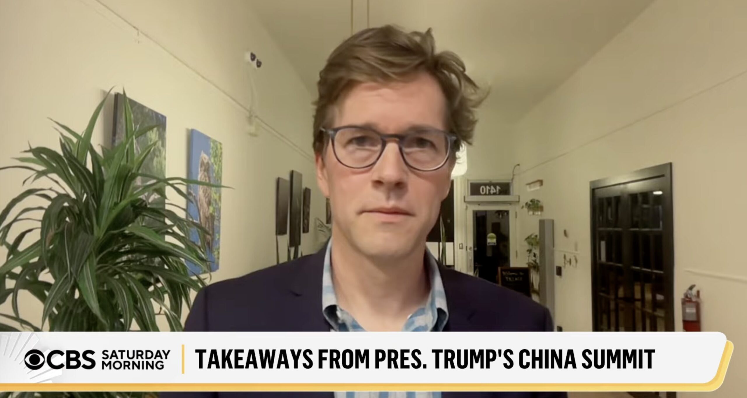 Man wearing glasses and a blazer stands in a hallway with plants and art on the walls; CBS Saturday Morning banner shows 'Takeaways from Pres. Trump's China Summit' at the bottom.