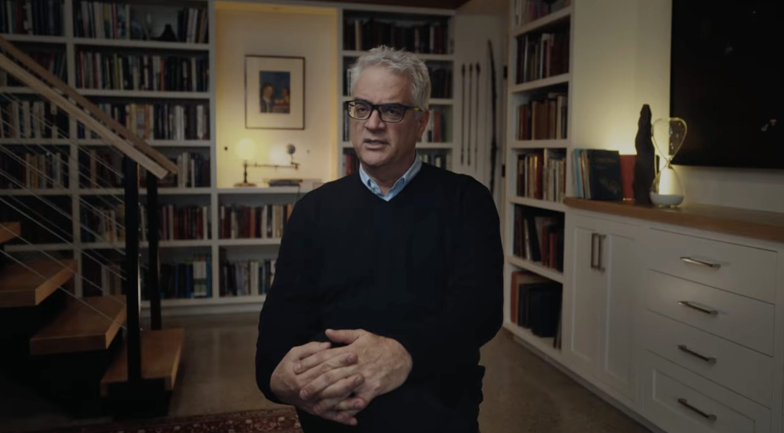 Man with gray hair and glasses seated in a home library, hands folded in his lap, facing forward.