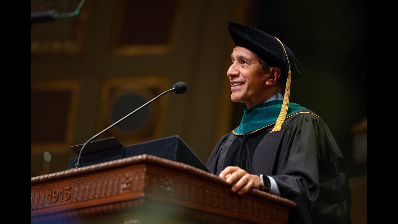 Smiling man in doctoral regalia delivers a speech at a wooden podium.