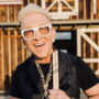 Joyful man with white hair and large white glasses, holding drumsticks outdoors with a wooden background.
