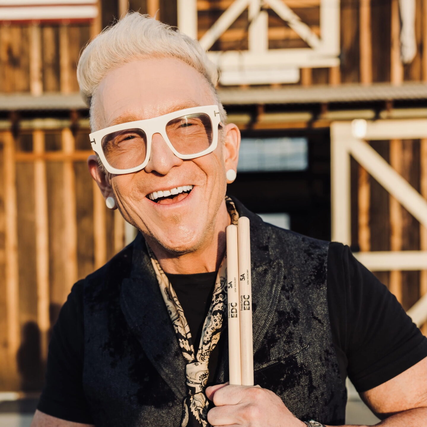 Joyful man with white hair and large white glasses, holding drumsticks outdoors with a wooden background.