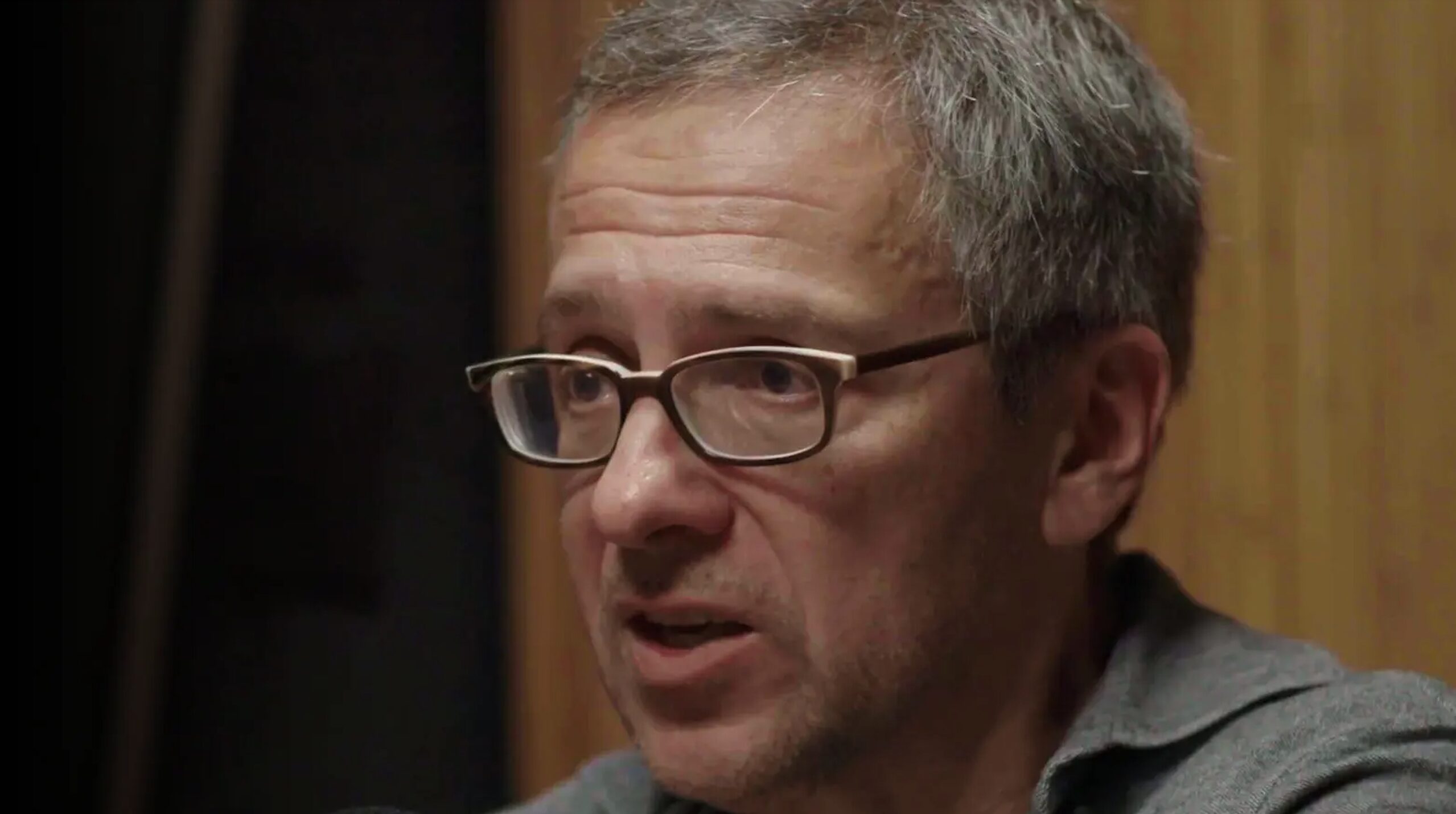 Close-up of a man with short gray hair and rectangular glasses speaking or mid-conversation.