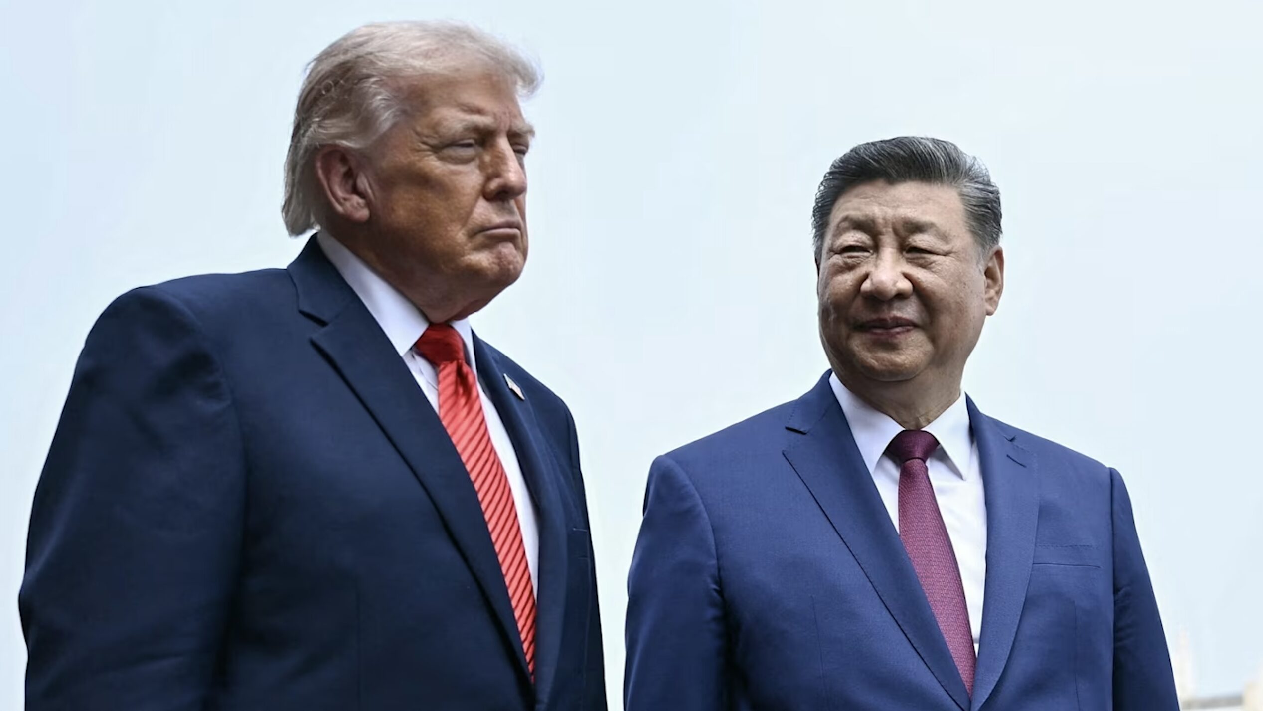Two men in navy suits stand side by side outdoors, looking serious; left wears a red striped tie and white shirt, right a lavender tie.