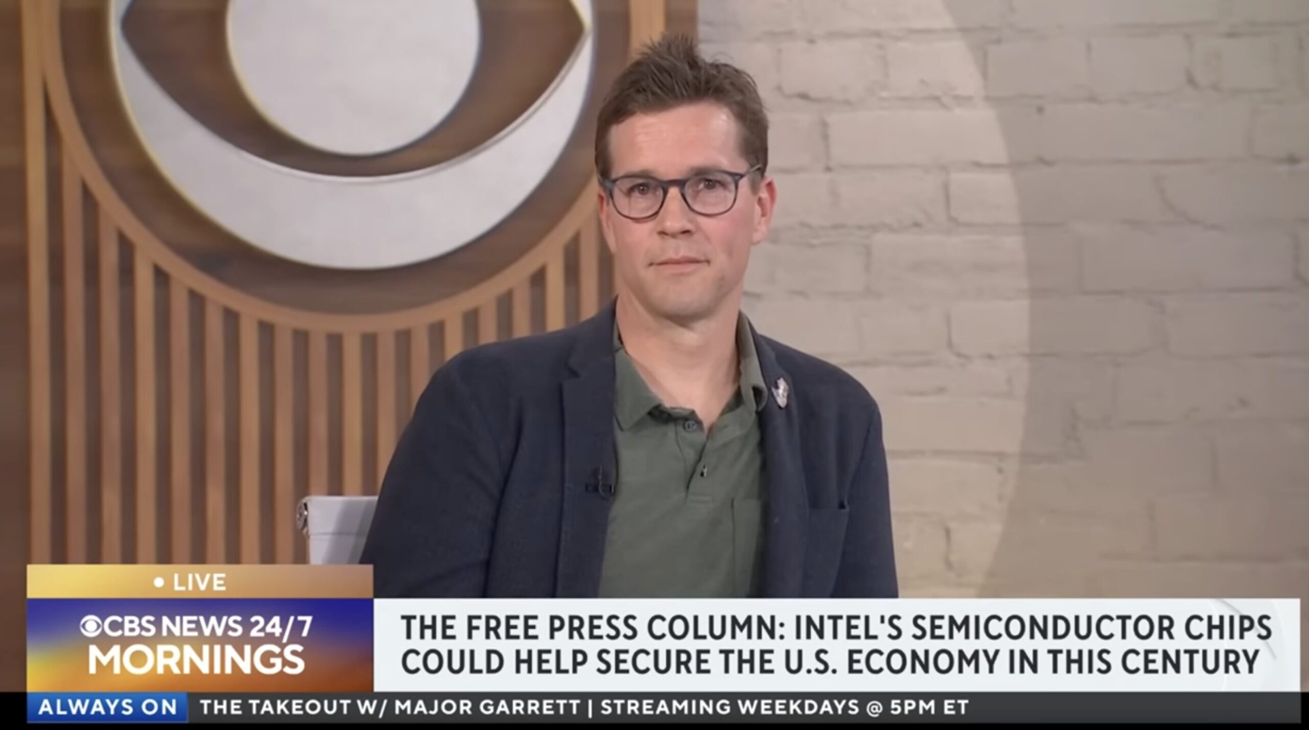 Male news host with glasses in blazer delivers a live segment about Intel semiconductors in a studio setting with a brick wall background and CBS banner.