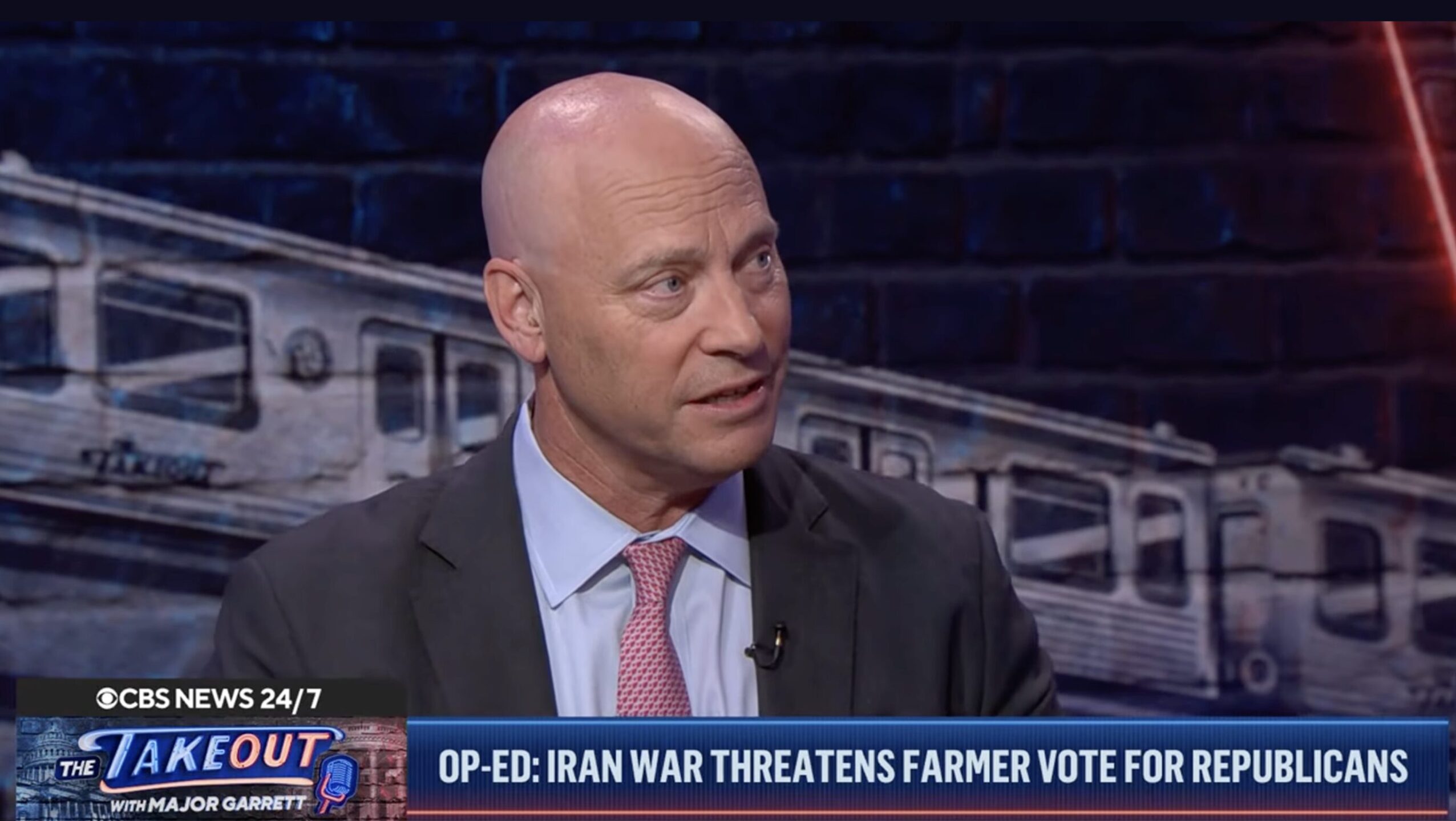 A man in a suit being interviewed on a TV news program with a blue ticker and the text 'OP-ED: IRAN WAR THREATS... FARMER VOTE FOR REPUBLICANS' in the lower third.