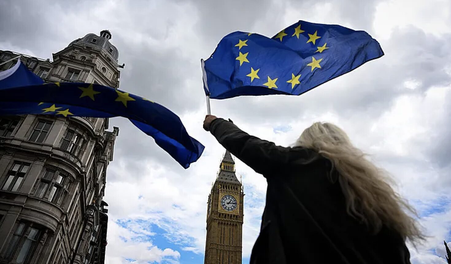 Person waving a blue European Union flag in front of Big Ben, London, under a cloudy sky.