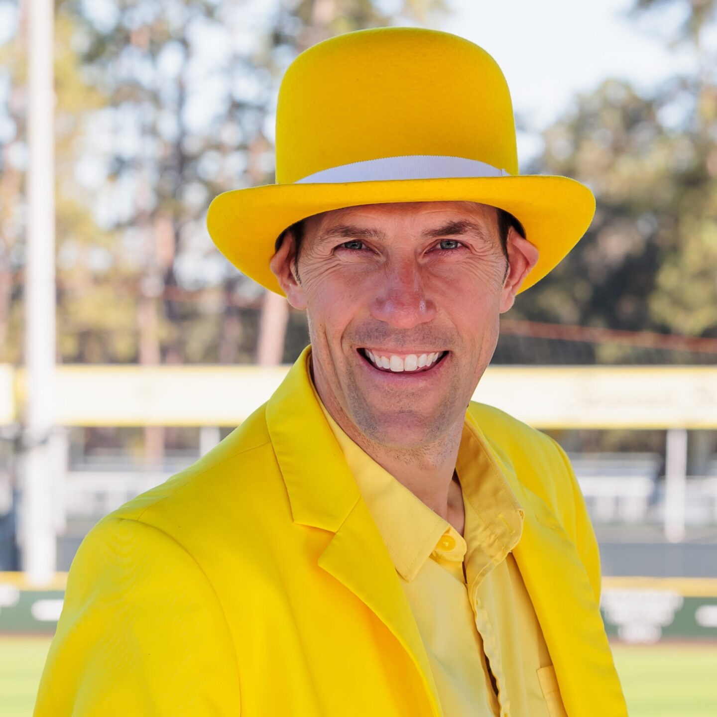 Smiling man in a bright yellow suit and hat, outdoors at a sunny sports venue.