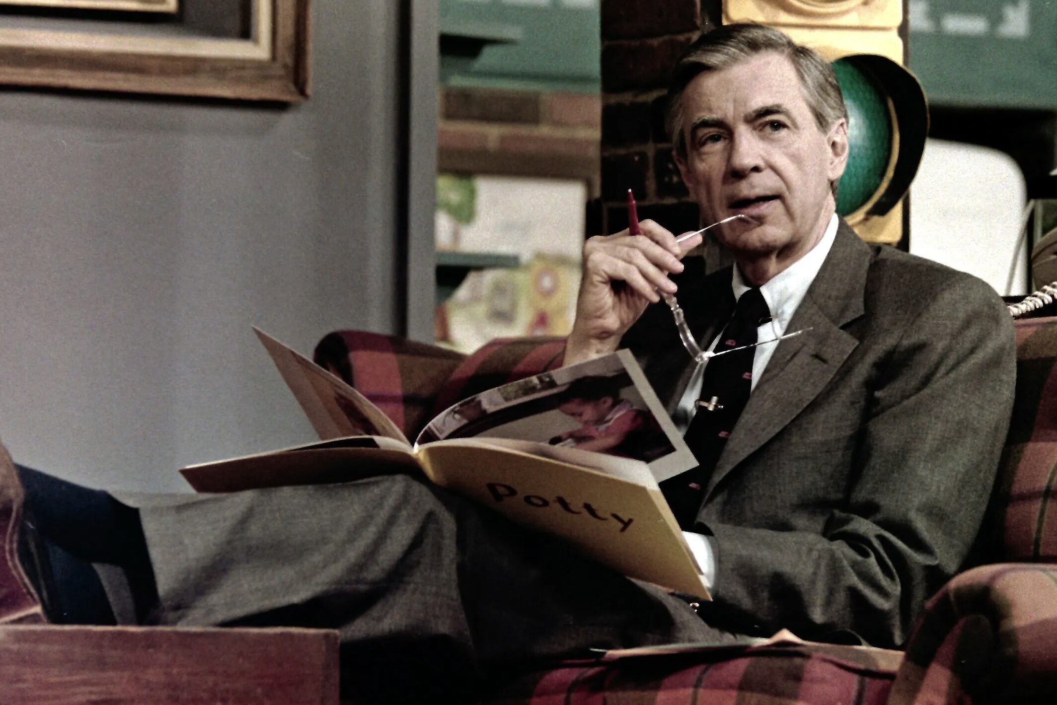 Older man in a gray suit reads an open, photo-filled book while holding his glasses.