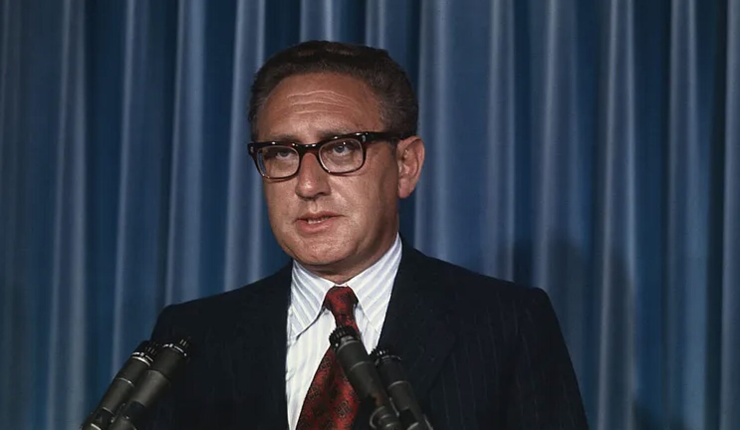 Man in a dark suit and red tie speaks at a podium with several microphones, blue curtain backdrop behind him.