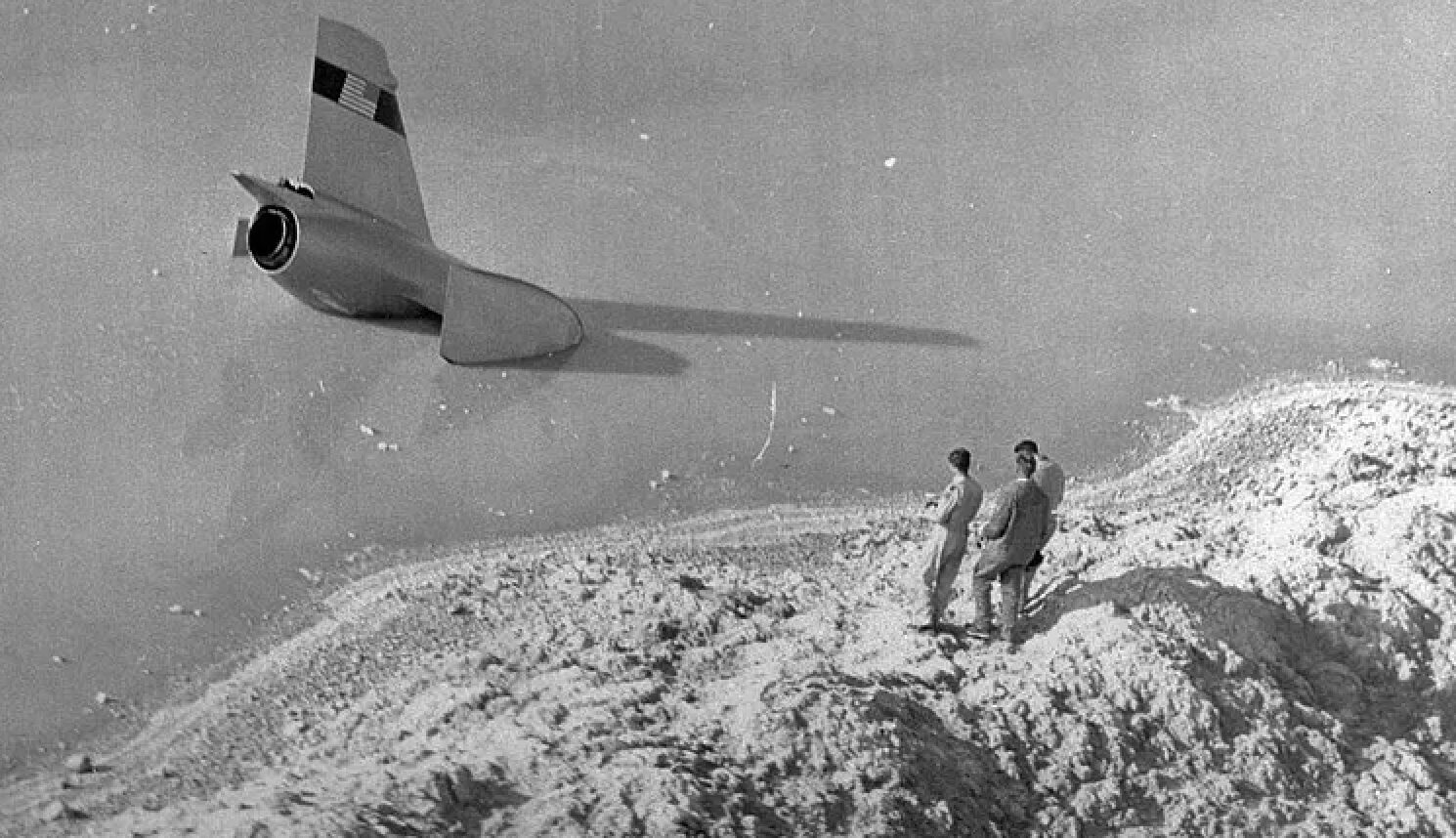 Two men stand on a rocky hilltop watching a jet fly low over a barren landscape.