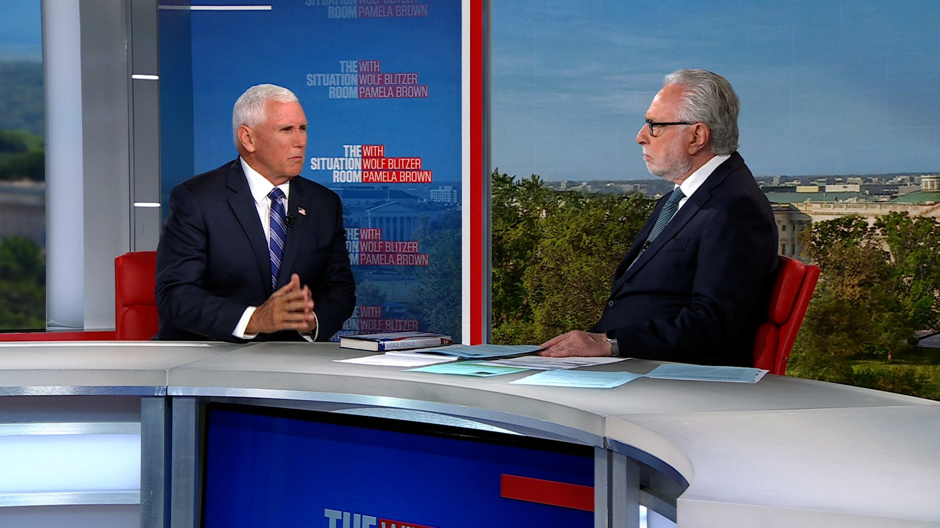 Two men in business suits sit at a news studio desk during a televised interview, with a blue backdrop bearing 'The Situation Room' graphics behind them, and papers on the desk.
