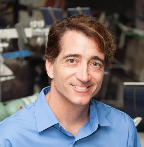 Smiling man with brown hair wearing a light blue shirt, in a busy lab or workshop setting behind him.